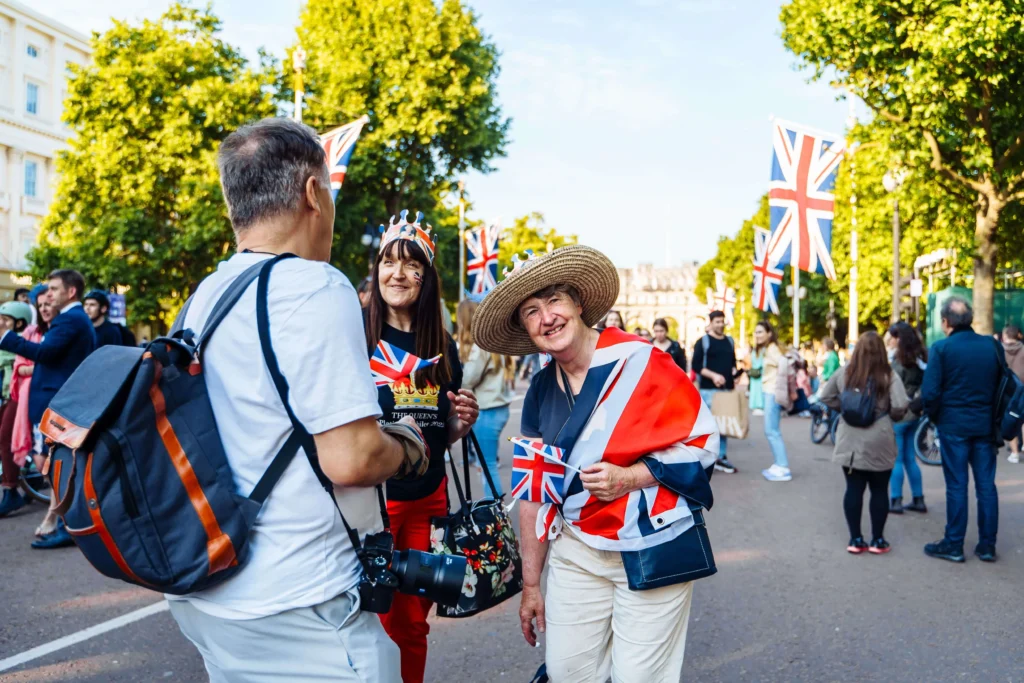 Three people celebrate outdoors with Union Jack flags, dressed festively, engaging cheerfully. Flags and trees line the sunny street, suggesting a UK event.