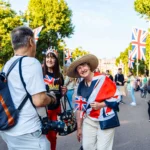 Three people celebrate outdoors with Union Jack flags, dressed festively, engaging cheerfully. Flags and trees line the sunny street, suggesting a UK event.